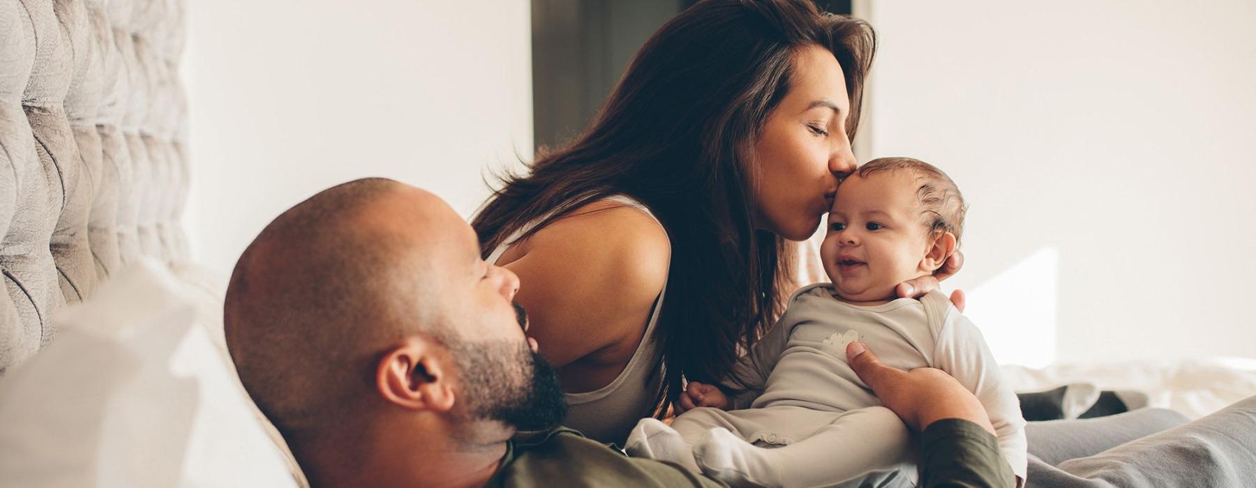 mother kisses her infant while father lies in bed and holds him